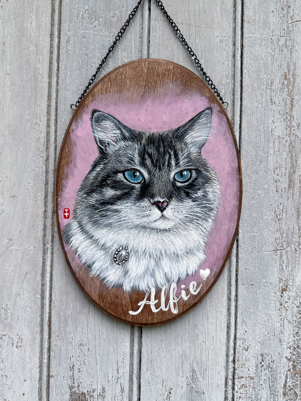 Hand-painted custom cat portrait on a walnut wooden oval plaque with black hanging chain, gray tabby cat with blue eyes on a pink background, name Alfie in white script