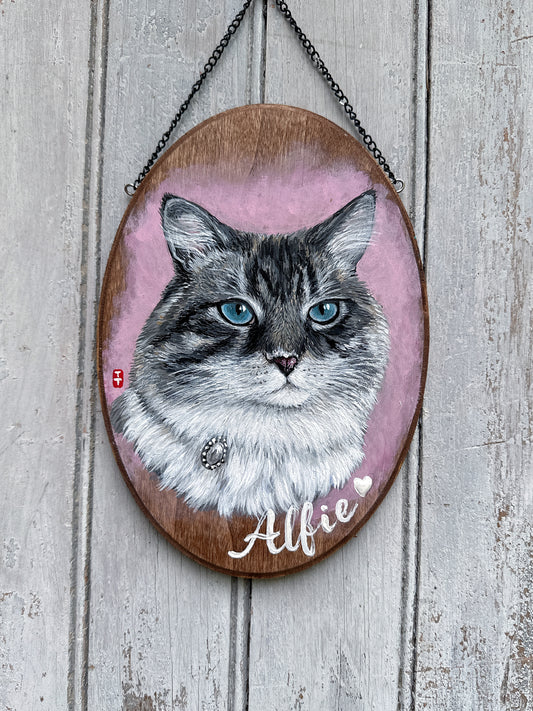 Hand-painted custom cat portrait on a walnut wooden oval plaque with black hanging chain, gray tabby cat with blue eyes on a pink background, name Alfie in white script