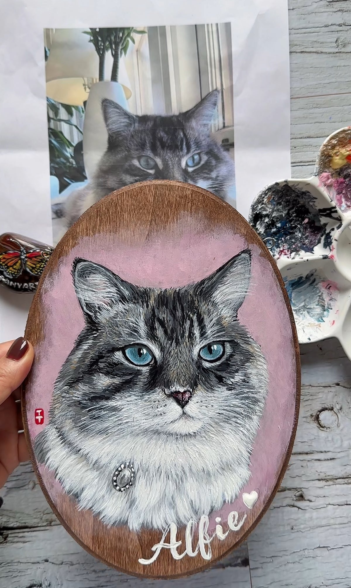 Artist holding the finished cat portrait plaque next to the customer's reference photo of the cat
